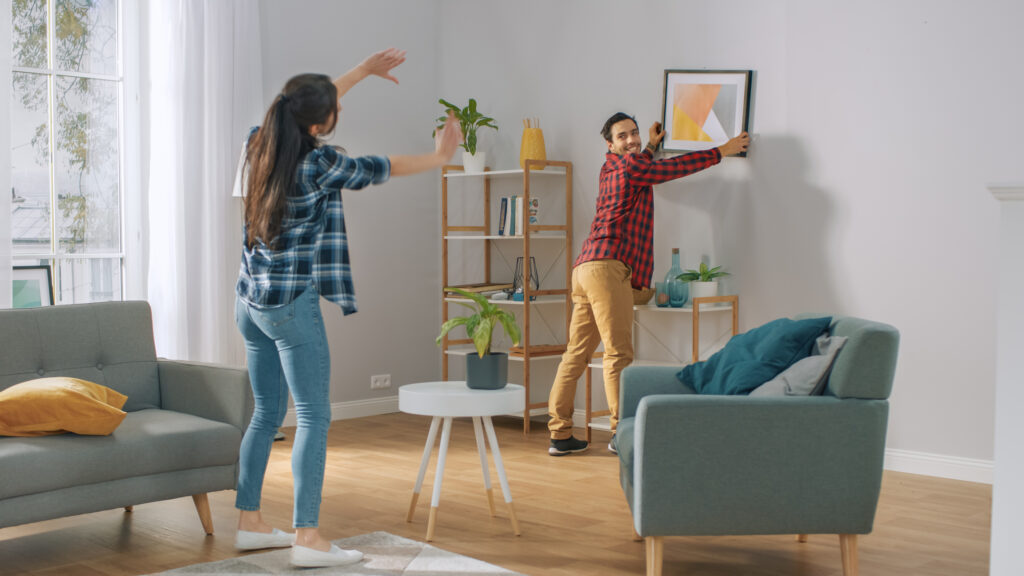 decorating on a budget. Happy Couple Hanging Picture on the Wall, Boyfriend Moves It, Girlfriend Tells Him when the Frame is Hanging Straight. Funny Moment in Young Couple's Life. Modern Stylish Apartment.