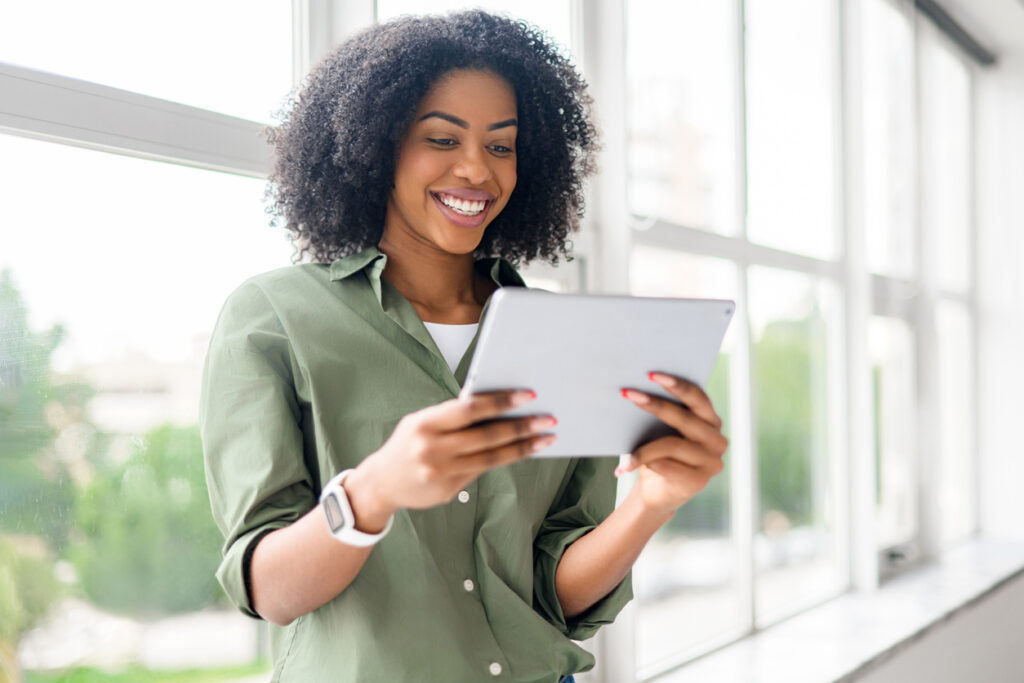 strategic about spending. An African-American woman with a beaming smile uses a tablet, engaging in a video call or browsing digital content, standing by a window that floods the room with natural light.